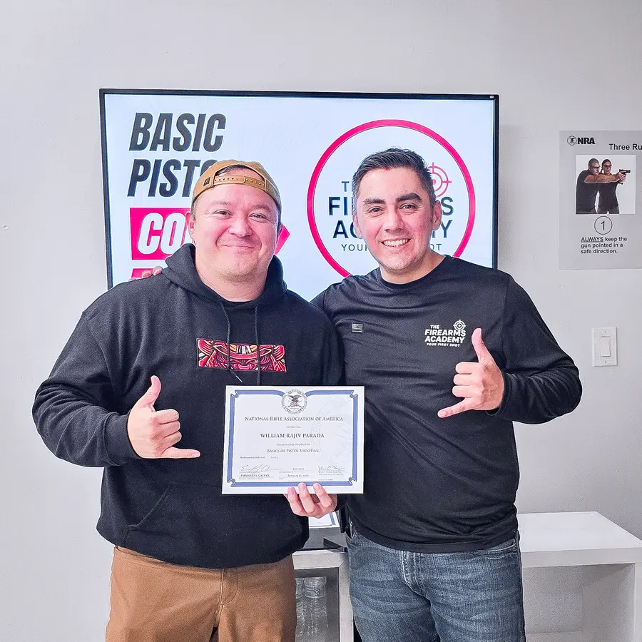 An instructor handing a student their NRA Basic Pistol course completion certificate, beside a Firearms Academy Basic Pistol course poster