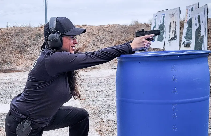A student kneeling behind a barrel used as cover, pistol extended over the top, shooting at targets on an outdoor range